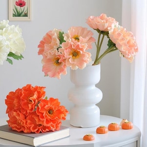 May include: A white vase holds peach-colored flowers with yellow centers. A bouquet of orange flowers sits on a book. Small orange pumpkin decorations are on a white table. A framed picture of a red flower hangs on the wall.