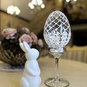 Peut inclure: Un lapin blanc en céramique est placé à côté d'un œuf décoratif en verre. L'œuf est enveloppé d'un filet de dentelle blanche et repose sur une tige en verre transparent. L'arrière-plan présente un arrangement floral flou et un éclairage doux.