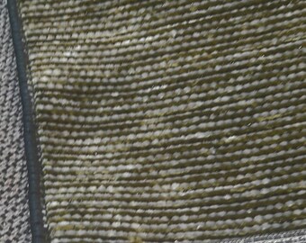 Puede incluir: Primer plano de una alfombra tejida verde y blanca con un borde de flecos. La alfombra tiene una superficie texturizada y un patrón sutil.