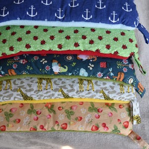 Könnte beinhalten: Ein Stapel bunter Stoffbeutel mit verschiedenen Mustern. Der oberste Beutel ist marineblau mit weißen Ankern. Weitere Beutel zeigen Marienkäfer, Giraffen und Erdbeeren. Jeder Beutel hat einen Reißverschluss und eine Stoffschlaufe.