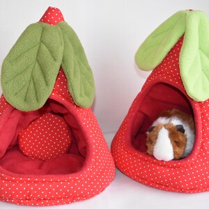 Könnte beinhalten: Zwei rote, erdbeerförmige Haustierbetten mit weißen Tupfen. Jedes Bett hat grüne Blätterakzente und einen roten Innenraum. Ein Bett enthält ein kleines, braun-weißes Meerschweinchen. Diese gemütlichen Betten sind für kleine Haustiere konzipiert.