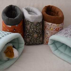 Könnte beinhalten: Eine Sammlung kleiner Haustierbetten in verschiedenen Mustern und Farben. Die Betten sind zylindrisch und oben offen, mit einem weichen, plüschigen Innenraum. Ein Bett enthält ein Meerschweinchen. Die Muster umfassen Tiere, Blätter und geometrische Formen.