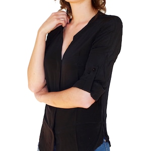 May include: Black button-down shirt with a V-neck collar and three-quarter sleeves. The shirt has a curved hemline and a small pocket on the left side. The sleeves are rolled up and secured with a button. The shirt is paired with blue jeans.