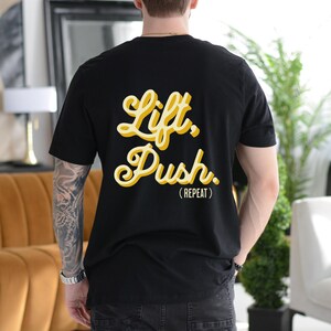 Könnte beinhalten: Schwarzes T-Shirt mit gelber Grafik, auf der "Lift, Push. (REPEAT)" steht.