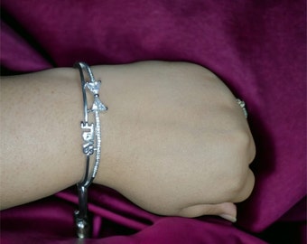 May include: Two silver-toned bracelets. One features a bow design with clear stones, and the other spells out "SMILE" in cursive. The bracelets are worn on a wrist, showcasing their delicate design and sparkle. The background is a deep purple.