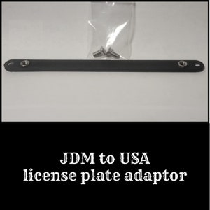 May include: A black license plate adaptor with two screws and nuts. The adaptor is a long, rectangular shape with holes at each end for mounting. The text "JDM to USA license plate adaptor" is at the bottom.