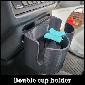 May include: A black double cup holder for a car. The cup holder is made of plastic and has a textured surface. The cup holder is designed to hold two cups or bottles.