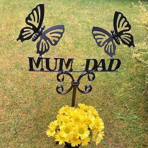 May include: Black metal garden memorial with two butterfly silhouettes and the words "MUM DAD" on a sign. A vase with yellow flowers is attached to the base.
