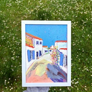 Op de afbeelding: Een ingelijst olieverfschilderij met een straatscène in mediterrane stijl met witte gebouwen, rode daken en blauwe luiken onder een helderblauwe lucht. Het kunstwerk bevat kleurrijke bloemaccenten en een getextureerd pad, alles in een eenvoudige witte lijst.
