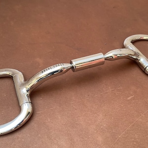 Op de afbeelding: Een gepolijste metalen paardenbit met een centrale roterende cilinder en D-vormige ringen. De bit heeft de tekst "ARSAL FORGE" gegraveerd op het gebogen gedeelte. De achtergrond is een bruin lederen oppervlak.