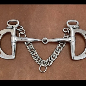 May include: A silver horse bit with a D-ring on each side and a chain connecting the rings. The bit is engraved with the words "ARSAL FORGE".