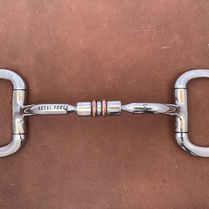 May include: A stainless steel horse bit with a D-ring design. The bit has a central mouthpiece with copper rings and the text "ARSAL FORCE" and "5" engraved on the sides. The bit is designed for equestrian use.