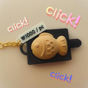 May include: A keychain featuring a tan, fish-shaped taiyaki charm set within a black rectangular frame. A small white rectangular tag attached to the gold-colored chain displays the black text ₩1000 / pc. The word click! appears in three locations.