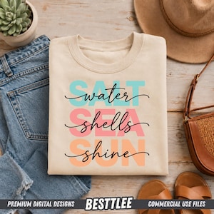May include: Cream-colored sweatshirt with the words "water, shells, shine" in a script font, with the words "water" in turquoise, "shells" in pink, and "shine" in orange. The sweatshirt is laid on top of blue jeans.