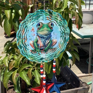 May include: A metal wind spinner with a patriotic frog design. The frog is wearing a red, white, and blue bandana and is surrounded by blue and green swirls. The spinner has three hanging stars in red, white, and blue.