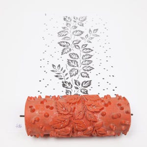 May include: An orange paint roller with a leaf design. The roller is used to create a repeating pattern on a wall or other surface. The roller is shown next to a sample of the pattern it creates.