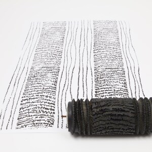 Op de afbeelding: Een zwarte textuurpatroonroller naast een wit papier met een zwart boomschorspatroon. De roller is gemaakt van een donker materiaal en heeft een cilindrische vorm. Het papier toont een herhalend patroon.