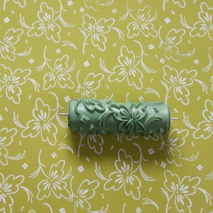 Op de afbeelding: Een groene verfroller met een bloemenpatroon. De roller ligt op een gele achtergrond met een wit bloemenpatroon.