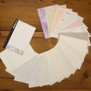 Op de afbeelding: Een verzameling diverse briefpapierartikelen, waaronder een notitieblok met de merknaam "Pelikan" en een verscheidenheid aan bedrukte en gelinieerde papieren. De papieren zijn in een waaier op een houten oppervlak gerangschikt.