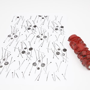 May include: A red patterned paint roller next to a white paper with a black ink pattern of lines and circles. The roller is cylindrical with a textured surface. The paper has a hand-drawn style.