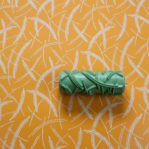 May include: A green paint roller with a textured design for creating a pattern on a wall. The roller is on a background of orange with white lines.