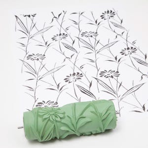 Könnte beinhalten: Eine grüne Farbrolle mit floralem Design, neben einem Papier mit einem schwarz-weißen Blumenmuster. Die Rolle hat ein detailliertes geprägtes Muster aus Blumen und Blättern. Das Papier zeigt ein sich wiederholendes Muster aus Blumen und Stielen.