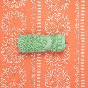 Könnte beinhalten: Eine hellgrüne Musterwalze mit einem detaillierten floralen und geometrischen Prägemuster, die horizontal auf einer leuchtend orangefarbenen Oberfläche mit einem weißen, sich wiederholenden Blumen- und Streifenmuster liegt.
