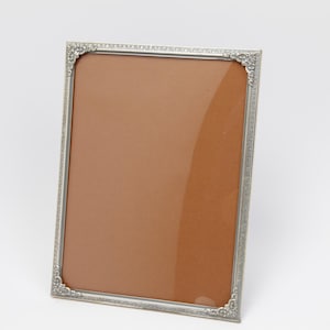 Op de afbeelding: Een rechthoekige zilverkleurige fotolijst met sierlijke hoekdetails. De lijst heeft een bruine achterkant en een heldere glazen voorkant. De lijst staat schuin, wat het ontwerp en de ruimte voor een foto of kunstwerk benadrukt.