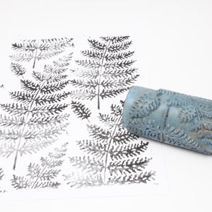 May include: A blue fern pattern roller beside a print of fern leaves in black and grey on white paper. The roller has a textured surface with a fern leaf design. The print shows multiple fern leaf impressions.