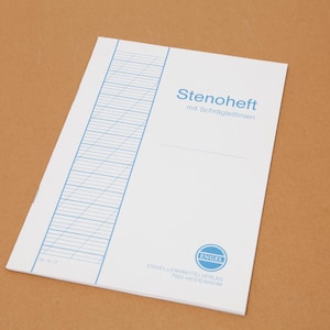 Può includere: Un quaderno bianco con linee diagonali blu e il testo tedesco "Stenoheft mit Schräglinien" è visualizzato su una superficie marrone. Il quaderno ha un logo blu e testo in basso.