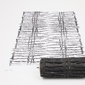 May include: A black textured roller with a wooden handle sits next to a black and white print. The print shows a grid-like pattern of horizontal and vertical lines, creating a textured effect. The roller is likely used for creating the print.