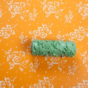 Op de afbeelding: Een groene verfroller met een bloemenpatroon. De roller bevindt zich op een feloranje achtergrond met een wit bloemenpatroon.