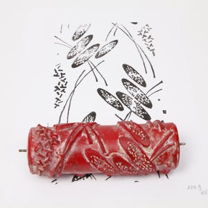 Könnte beinhalten: Eine rote Musterwalze mit einer erhabenen Textur, die ein sich wiederholendes Blattmotiv erzeugt. Die Walze liegt auf einem weißen Blatt Papier mit einem schwarzen Abdruck desselben Blattmusters, wobei der Text 124 g KHH zu sehen ist.