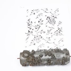 May include: A vintage metal pattern roller with a floral design, creating a repeating pattern on a white surface. The roller is grey and has a detailed, textured surface. The floral pattern includes flowers, leaves, and geometric shapes.