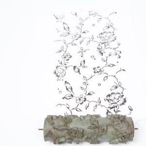 May include: A decorative floral pattern roller with a detailed design, next to a white paper with a black floral pattern. The roller is a muted green color and appears to be made of metal. The floral pattern on the paper is black and white.