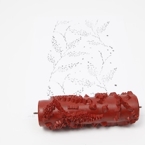 May include: A red patterned paint roller with a textured design, used to create a floral pattern on a white surface. The roller is cylindrical and has a metal rod through the centre.