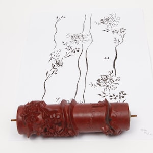 以下が含まれることがあります： 浮き彫りの花柄が施された円筒形の赤いペイントローラー。ローラーは白い紙の下部に配置されており、紙には同じ花柄が黒インクで印刷され、垂直な茎とバラが描かれています。
