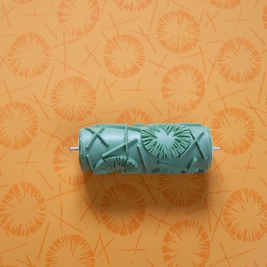 Op de afbeelding: Groene verfroller met een geometrisch patroon om een ​​herhalend ontwerp op muren of andere oppervlakken te creëren. De roller bevindt zich op een gele achtergrond met een herhalend patroon van oranje cirkels.