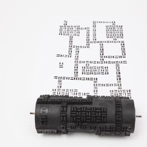 Könnte beinhalten: Ein schwarzer Rollstempel mit geometrischem Muster, der ein sich wiederholendes Muster auf einer weißen Oberfläche erzeugt. Der Stempel ist zylindrisch mit strukturierter Oberfläche und Metallstiften an jeder Seite. Das gestempelte Muster ist schwarz.