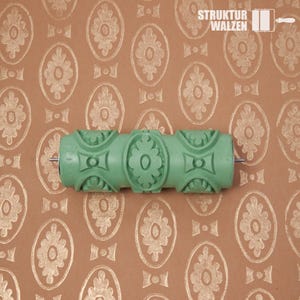 May include: A sage green patterned paint roller featuring a repeating oval and floral motif. The roller is positioned over a brown surface displaying the same stamped pattern in gold ink. The text STRUKTUR WALZEN is visible in the top right corner.
