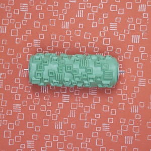 Op de afbeelding: Een getextureerde, mintgroene verfroller met een geometrisch patroon, geplaatst tegen een koraalrode achtergrond met een herhalend wit vierkant en lijnenontwerp. De roller is ontworpen voor het creëren van patrooneffecten op muren of andere oppervlakken.