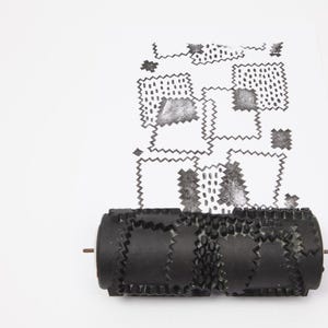 Può includere: Un timbro a rullo nero con motivi è mostrato con un foglio di carta che mostra un motivo ripetuto di forme timbrate. Il timbro ha una serie di motivi strutturati e ripetuti. Il design timbrato include quadrati, zig-zag e punti.