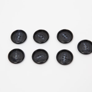 Può includere: Sette bottoni neri con finitura lucida sono disposti su una superficie bianca. Ogni bottone ha quattro fori e un sottile motivo marmorizzato nei toni del grigio e del bianco, che aggiunge un tocco di interesse visivo al design classico.