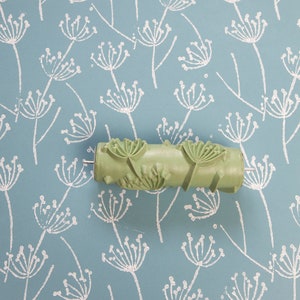 May include: A green paint roller with a design of stylised flowers. The roller is on a blue background with a white pattern of stylised flowers.