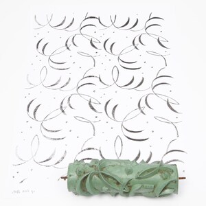 Op de afbeelding: Een groene verfroller met patroon ligt onder een wit oppervlak met een herhalend zwart wervelontwerp. De roller heeft een bloemenpatroon en een metalen staaf. Het ontwerp op het witte oppervlak is een herhalend patroon van zwarte wervelingen en stippen.