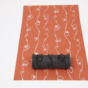 May include: A black pattern roller rests on a terracotta-colored paper with a white, repeating design. The roller has a textured surface with a floral-like pattern. The paper has a repeating white design of curved lines and oval shapes.
