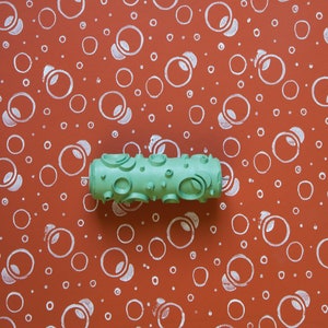 May include: A green paint roller with a textured surface designed to create a pattern of circles. The roller is on a red background with white circles.