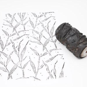 May include: A black, textured roller stamp next to a white paper with a black leaf pattern. The stamp has a cylindrical shape with a detailed, carved design. The leaf pattern is a repeating design of long, thin leaves.