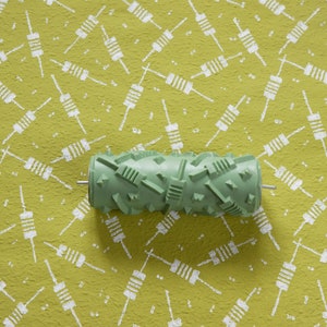 May include: A green textured paint roller with a handle and a metal rod. The roller is on a green surface with a white pattern of small, repeated shapes.