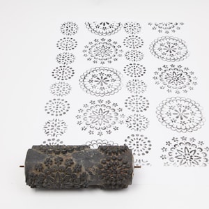May include: A dark, textured roller with a floral pattern is shown next to a white surface with a repeating floral design. The roller is used for creating patterns on surfaces. The floral design is in shades of grey.